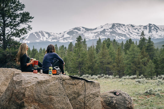 Two women camping on a rock with portable power station, pine trees, snowy mountains in background