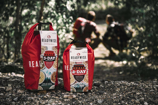 ReadyWise adventure meals 3-day and 2-day kits on forest ground, hikers in background