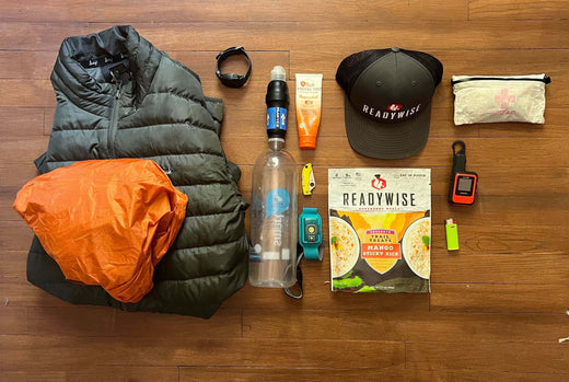 Flat lay of hiking essentials including jacket, ReadyWise hat, meal pouch, water bottle, gear on wood floor