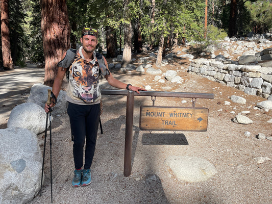 Hiker with trekking poles at Mount Whitney Trail sign in forest, rocky path visible